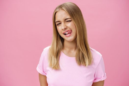 Playful flirty attractive young girl with fair straight hair in t-shirt winking at camera smiling and licking upper lip with tongue feeling enthusiastic and carefree against pink background. Emotions concept