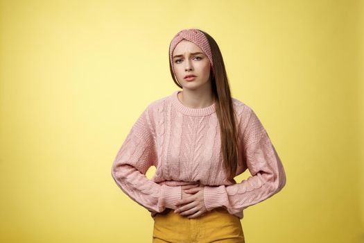 Girl suffering pain in stomach, having painful periods, feeling unwell touching belly grimacing displeased and troubled, having cramps, stooping upset of stomachahe against yellow background.