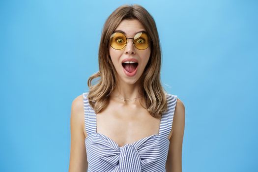 Waist-up shot of excited surprised and emotive charismatic caucasian woman in trendy sunglasses opening mouth from amazement and joy gazing at camera impressed and astonished over blue background. Lifestyle.