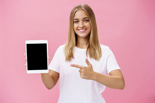Girl recommends use only this digital tablet. Charismatic young female with fair hair tanned skin and no makeup in white t-shirt pointing at gadget screen and smiling friendly at camera over pink wall. Technology and advertisement concept