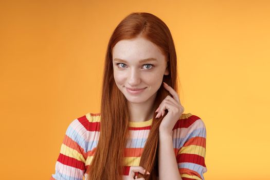 Romantic flirty shy attractive redhead girl 20s touching hair strand smiling silly modest glancing camera coquettish making lovely glances wanna seduce guy expressing sympathy, orange background.