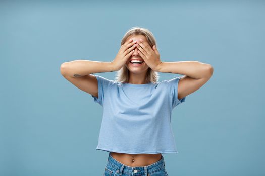 Hide if you can. Portrait of carefree and happy good-looking playful girl with blonde hair smiling broadly while counting ten and playing hide-and-seek with closed eyes over palms near blue wall.