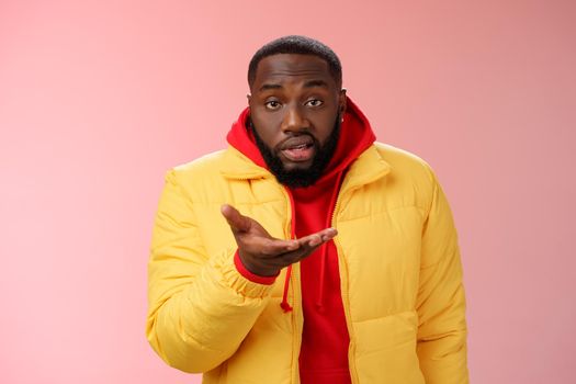 Bothered rude ignorant african-american bearded man pointing palm camera look dumb perplexed, standing confused cannot get clue what happening, wearing yellow jacket, pink background.