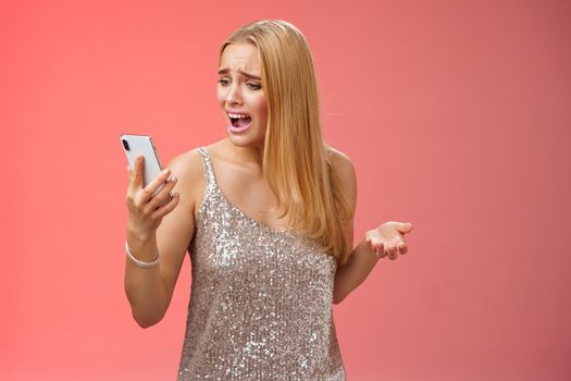 Troubled concerned arrogant young blond woman complaining yelling smartphone cannot call friend no signal holding smartphone look mobile display pissed moody arguging, red background.