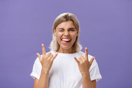 Gonna rock. Portrait of joyful happy good-looking stylish woman with blond medium haircut winking smiling and sticking out tongue while showing rock-n-roll gesture with both hands over purple wall.