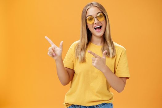 Click her watch my videos. Happy confident and outgoing charming young girl with straight hair in sunglasses and yellow t-shirt pointing at upper left corner promoting product with joyful smile.