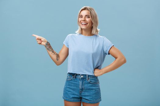 Girl proudly shows place where she works holding hand on waist in confident pose smiling and laughing happily pointing left with tattooed arm standing over blue background carefree. Emotions and advertising concept