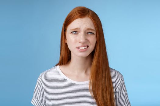 Displeased young awkward redhead girl cringe full disbelief smirking frowning confused look questioned doubtful hearing nuisance dumb story standing blue background uncertain. Copy space