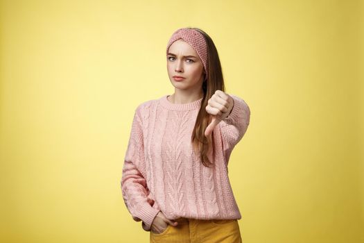 Not cool. Frustrated displeased and disagree cute young woman wearing knitted sweater frowning showing thumb-down smirking judgemental, expressing disapproval of unwell situation over yellow wall.
