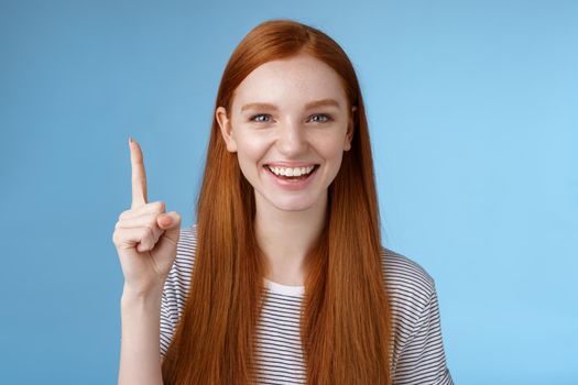 Got excellent idea pick for you. Attractive happy friendly-looking helpful redhead female shop assistant showing gorgeous dress customer pointing up index finger smiling glad help, blue background.