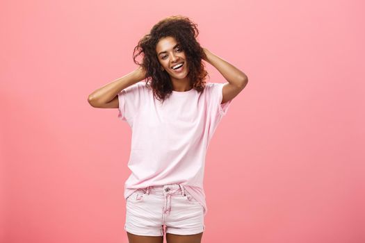 Girl enjoying nice weather chilling making photos for social network touching curly hair gently and carefree smiling broadly at camera standing over pink background happy and chill. Emotions and people concept