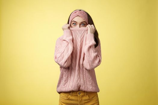 Girl hiding in collar wearing knitted sweater over nose popping eyes, staring bugged eyes at camera, hiding face reacting amazed and shocked, feeling scared, standing in stupor over yellow background.