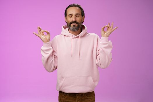 Peaceful charming hipster old man wearing cool pink hoodie close eyes breathing practice release stress meditating standing relaxed happy yoga meditation pose nirvana gesture, purple background.