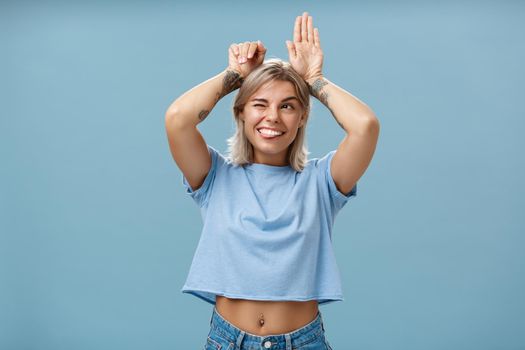 Lifestyle. Studio shot of entertained carefree and emotive happy charming woman with tattoos on arms acting like bunny with palms on head winking joyfully smiling and sticking out tongue over blue wall.