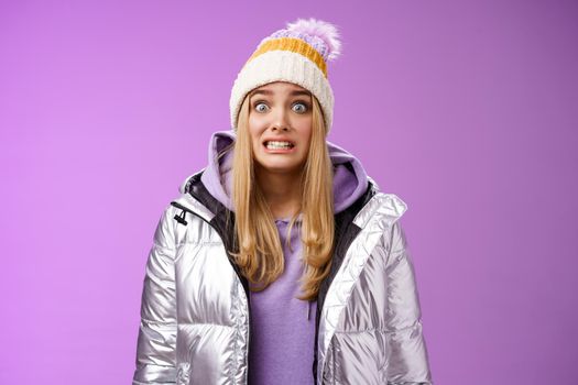Awkward worried cute timid blond girl in silver jacket hoodie winter hat clench teeth popping eyes camera ooops make mistake standing nervous someone notice, purple background. Copy space