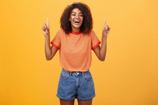 Woman feeling amused and entertained. Portrait of happy carefree stylish African-American girl with afro hairstyle laughing out loud joyfully pointing up with raised arms over orange wall. Emotions concept