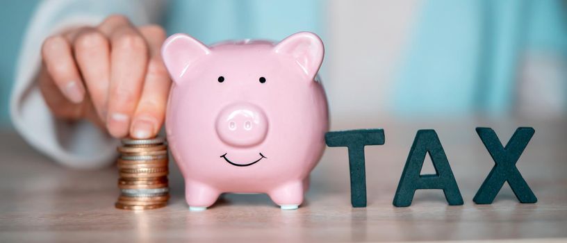 The hand of a young girl puts letters on the table with the words tax next to a piggy bank on the table at her workplace, close up view.