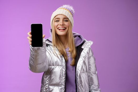 Sassy delighted cheerful blond woman recommend use app edit perfect pictures holding smartphone showing mobile phone display proudly satisfactory smile camera, standing purple background.