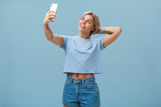 New post in my blog. Stylish feminine and sociable good-looking young female posing for selfie with pulled arm and smartphone sticking out tongue while making faces at device screen over blue wall.