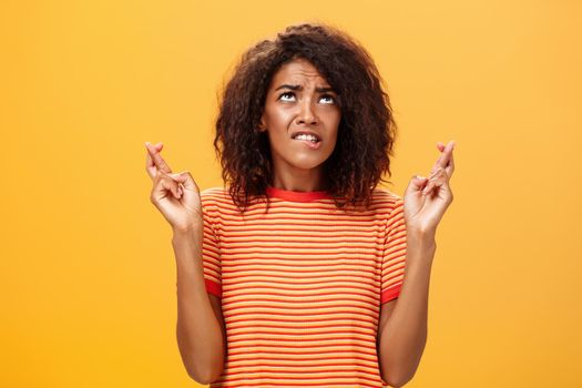 Waist-up shot of hopeful concerned and anxious charming dark-skinned woman with afro hairstyle in trendy striped t-shirt biting lip nervously looking up crossing fingers for good luck while praying. Lifestyle.