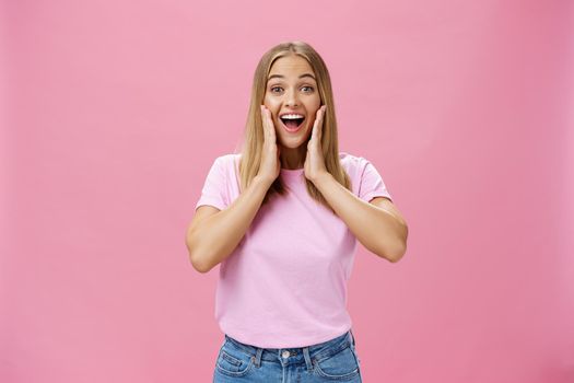 Delighted and amazed happy girlfriend in t-shirt and jeans smiling broadly with opened mouth amused touching cheeks surprised and satisfied reacting to positive surprise over pink background. Copy space