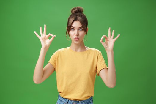 Portrait of silly cute and beautiful caucasian woman with combed hair in round earrings and yellow t-shirt showing okay, no problem gesture folding lips, reaffirming boss job goes well over green wall.