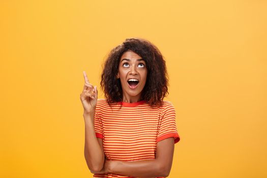 Waist-up shot of interested curious good-looking dark-skinned female in striped t-shirt talking asking question about curious star looking and pointing up with joy over orange background. Copy space