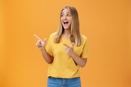 Amused enthusiastic and charismatic girl with blond hair and tanned pure skin without makeup pointing and looking at upper left corner with joyful happy smile over orange background. Body language concept
