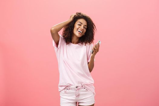 Portrait of carefree stylish modern dark-skinned young girl using smartphone hanging around joyfully touching hair and gazing with broad smile at camera holding cellphone posing against pink wall. Technology concept