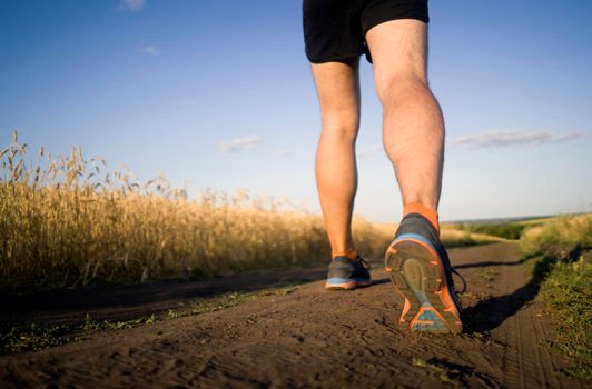 Athlete runner legs running on a close-up of a trail across a field on a shoe. Fitness and warm-up, young man with sun effect in the background.
