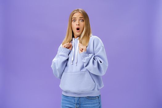 Woman scared of sudden loud noise in dark gasping looking shocked and concerned tilting backwards with frightened look pressing clenched fists to body from fear, wearing warm hoodie over purple wall. Emotions concept
