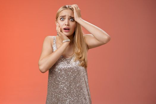Shocked frightened insecure panicking woman blond hairstyle in silver dress touch head pop eyes afraid scared seing terrifying crime standing stupor speechless gasping shook, red background.