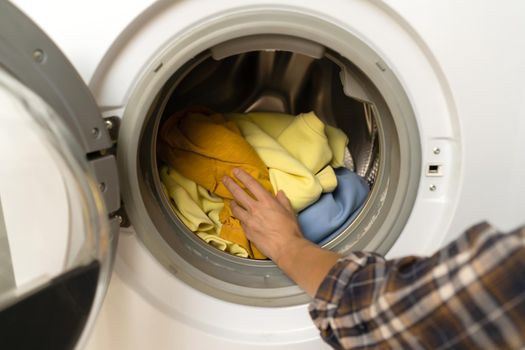 A girl folds bright clothes into a new washing machine in the bathroom at home, a woman washes things and spin them out with detergent in the laundry close-up, top view.