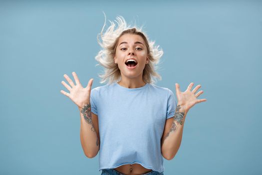 Creative happy and playful beautiful blond girl with tattoos on arms raising palms up opening mouth facing wind while hair strands flicking on air jumping having fun over blue background. Emotions concept