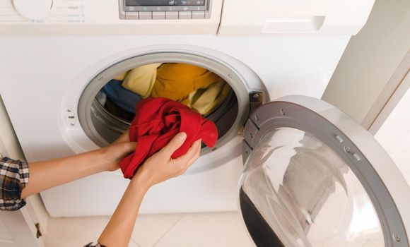 A girl folds bright clothes into a new washing machine in the bathroom at home, a woman washes things and spin them out with detergent in the laundry close-up, top view.