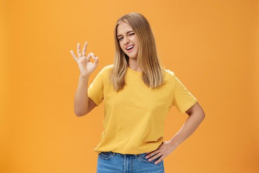 Confident and skillful female student assuring teacher she do great job holding hand on waist winking at camera with self-assured look and showing okay gesture assuring everything ok over orange wall. Lifestyle.