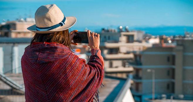 A young girl in a burgundy poncho and a hat travels and takes a photo with a cityscape on a sunny day.