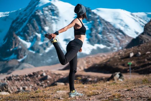 A young sports girl is warming up on a background of snow-capped mountains on a sunny day, a woman does exercises, trains and meditates in a picturesque mountainous area.
