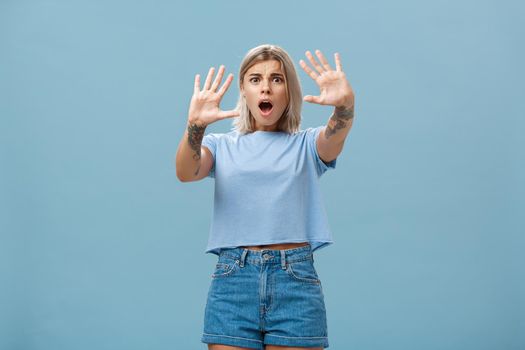 Woman terrified screaming and asking stop. Portrait of shocked panicking troubled blonde female in denim shorts and casual t-shirt pulling hands in no gesture dropping jaw and frowning. Copy space