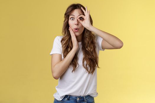 No way. Surprised impressed young funny attractive girl long chestnut hair fold lips admiration wow sound touch cheek astonished intrigued look through ok ring sign like monocle yellow background.