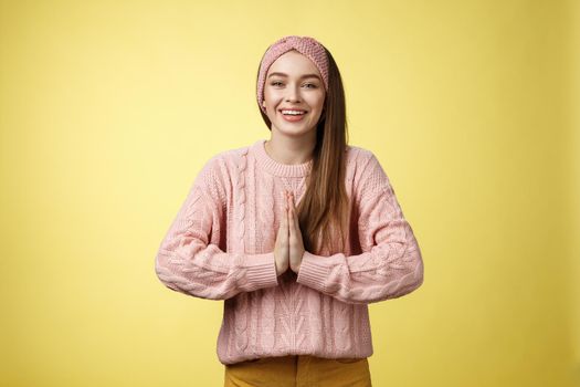 Please do me favour. Charming young female student wearing knitted sweater, headband pressing palms together in pray begging, asking for help smiling thankful, grateful for effort over yellow wall.