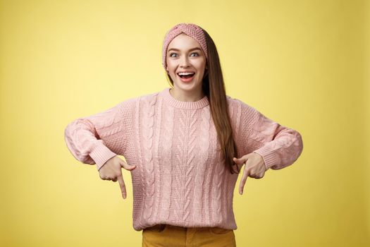 Lifestyle. Euphoric attractive young trendy girl in knitted band wearing sweater grinning excited enthusiastic pointing down thrilled of awesome promotion standing amused and overwhelmed over yellow background.