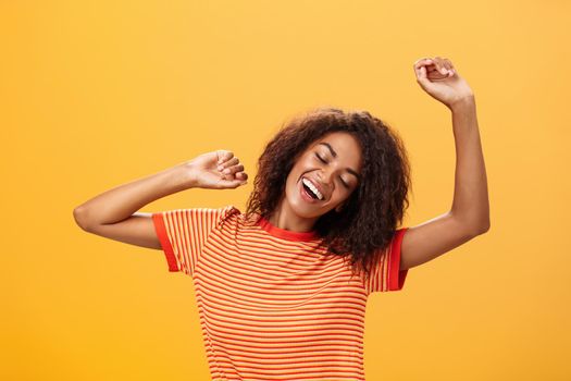 Waist-up shot of relaxed and relieved happy african american young female student in striped t-shirt stretching arms up closing eyes from delight and smiling having great nap over orange background. Lifestyle.