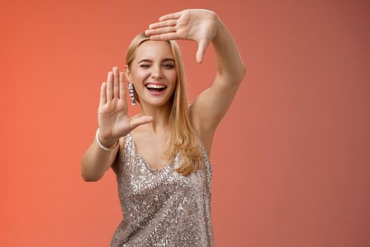 Sassy good-looking creative european blond woman in silver glittering dress winking cheeky smiling confident search inspiration around make frame hand search location take cool shot, red background.