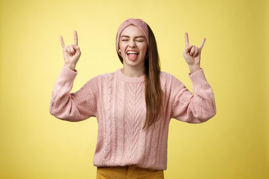 Lifestyle. Cute heavy metal lover showing rock roll symbol sticking tongue amused and happy fooling around listening favourite music posing excited and pleased against yellow background in knitted outfit.