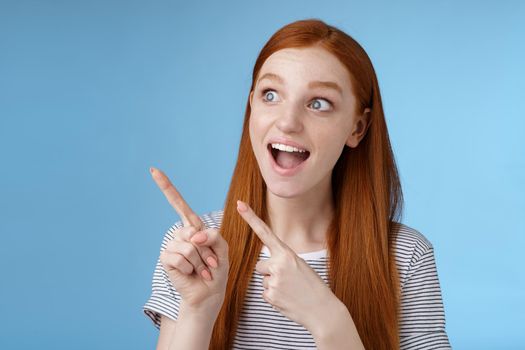 Lifestyle. Amused adorable redhead european girl say wow stare gasping astonished pointing looking upper left corner excited smiling drop jaw amazed standing thrilled blue background excitement concept.
