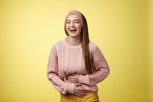 Amused and entertained charming young european female student laughing till belly hurts from giggle smiling holding palms on stomach, having fun enjoying watching funny comedy over yellow wall.