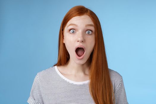 Wow so cool. Impressed speechless amused attractive redhead girl blue eyes freckles open mouth wide omg drop jaw astonished express surprise amazement, standing blue background. Copy space