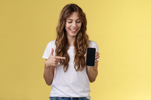 Intrigued charismatic happy young female blogger show what photo app use edit social media profile hold smartphone looking pointing index finger mobile phone blank screen smiling amused. Lifestyle.
