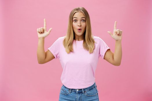 Amused and impressed attractive common european woman with fair hair and tanned skin raising index fingers pointing up amazed folding lips from interest and astonishment over pink background. Copy space